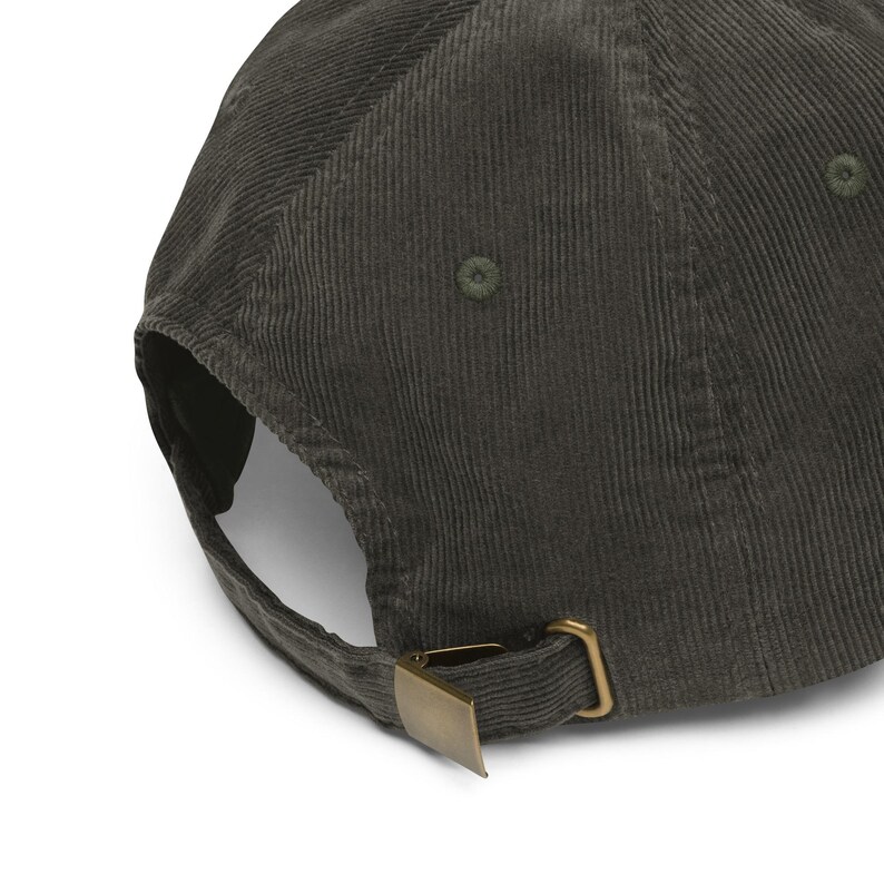 May include: A dark olive green corduroy baseball cap. The cap features a curved brim, a brass-colored metal clasp, and a fabric-covered button on top. The corduroy fabric has a vertical ribbed texture.
