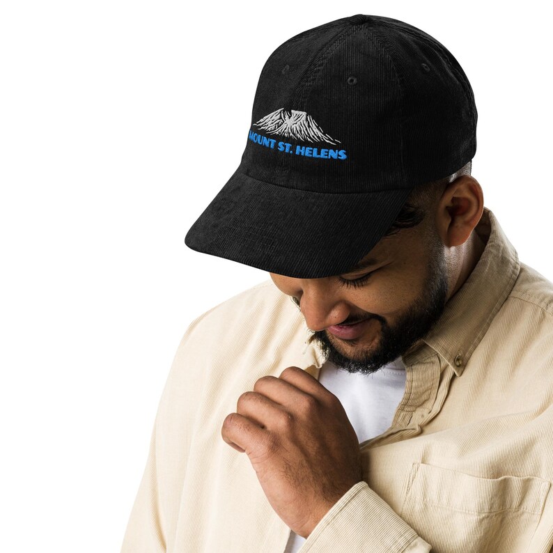 May include: Black corduroy baseball cap featuring an embroidered design of Mount St. Helens in white and blue. The text "MOUNT ST. HELENS" is arched above the mountain graphic. The cap has a curved brim and a structured fit.
