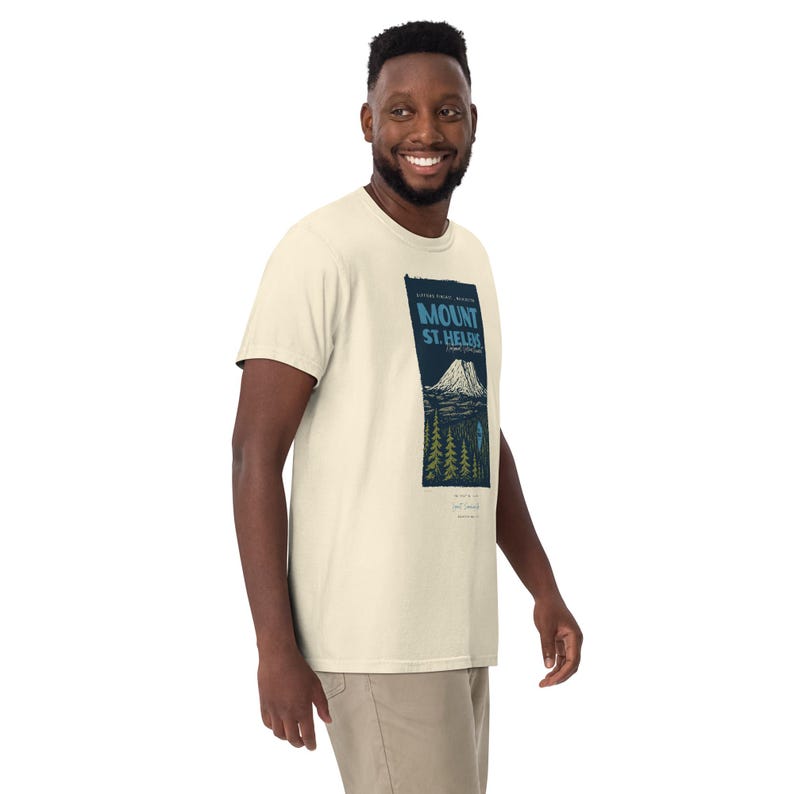 May include: Cream-colored t-shirt featuring a rectangular graphic with the text "MOUNT ST. HELENS" in blue and white. The graphic includes a mountain illustration and trees. The shirt is made of a soft material.
