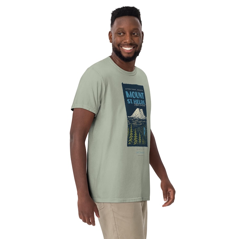 May include: Sage green t-shirt featuring a rectangular graphic with the text "MOUNT ST. HELENS" above a mountain illustration. The graphic also includes trees and a small figure. The shirt is made of a soft material.