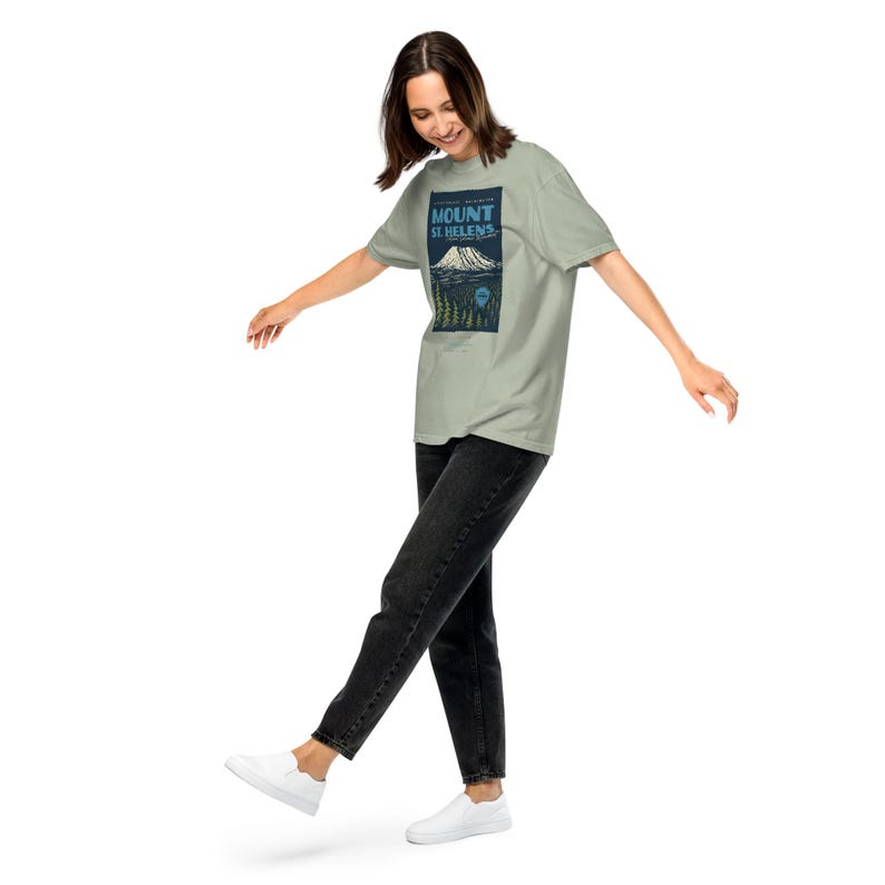 May include: A light green t-shirt featuring a graphic of Mount St. Helens. The design includes the mountain, trees, and text. The person is wearing black jeans and white shoes.