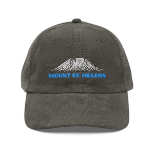 May include: Olive green corduroy baseball cap with a curved brim. The cap features an embroidered design of a mountain and the text "MOUNT ST. HELENS" in blue. The cap has a structured crown and an adjustable strap.