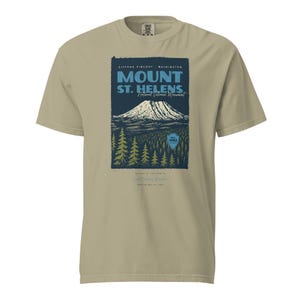 May include: A tan t-shirt featuring a graphic of Mount St. Helens National Volcanic Monument. The design includes the mountain, trees, and text that reads "Mount St. Helens" and "Gifford Pinchot, Washington."