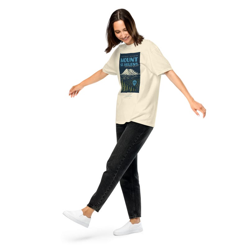 May include: Cream-colored t-shirt featuring a graphic of Mount St. Helens with text. The design includes a mountain, trees, and the words "Mount St. Helens" in a rectangular frame. The person is wearing black jeans and white shoes.