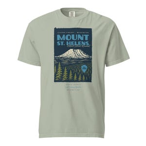 May include: A light green t-shirt featuring a graphic of Mount St. Helens National Volcanic Monument. The design includes a mountain, trees, and text that reads "Mount St. Helens" and "Gifford Pinchot, Washington."
