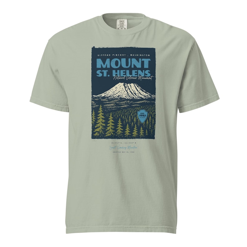 May include: A light green t-shirt featuring a graphic of Mount St. Helens National Volcanic Monument. The design includes a mountain, trees, and text that reads "Mount St. Helens" and "Gifford Pinchot, Washington."