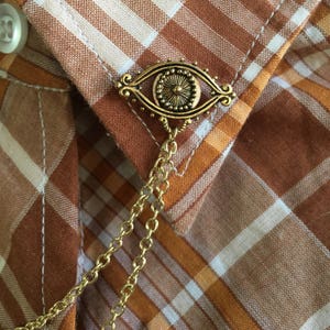 May include: A gold eye charm with a chain attached. The charm is on a brown and white plaid shirt.
