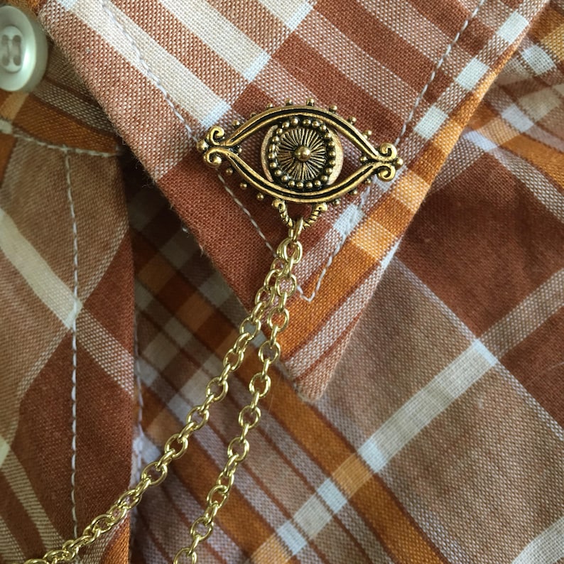 May include: A gold eye charm with a chain attached. The charm is on a brown and white plaid shirt.