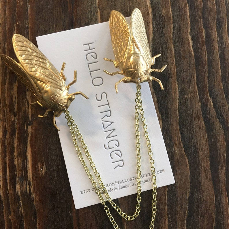 May include: Two gold cicada charms on a gold chain. The charms are detailed and realistic. The chain is attached to a white card with the text "HELLO STRANGER" and the Etsy shop name.