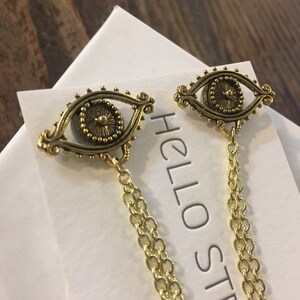 May include: Gold tone earrings with a detailed eye design and a dangling chain. The earrings are on a white card with the text "HELLO ST" visible.
