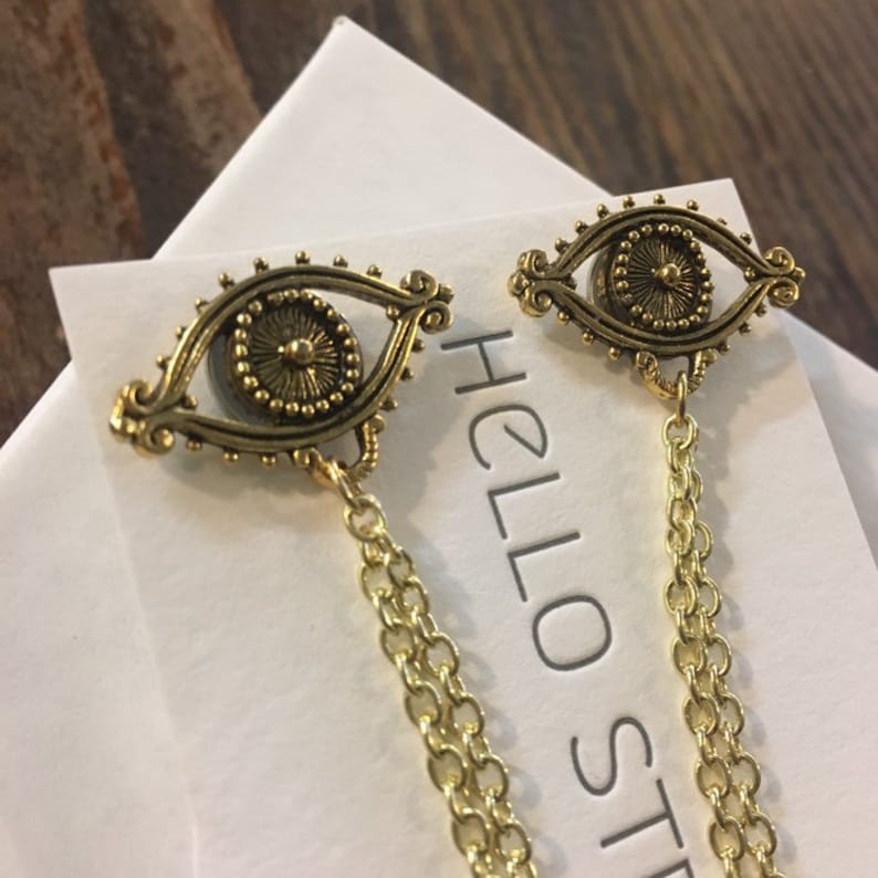 May include: Gold tone earrings with a detailed eye design and a dangling chain. The earrings are on a white card with the text "HELLO ST" visible.