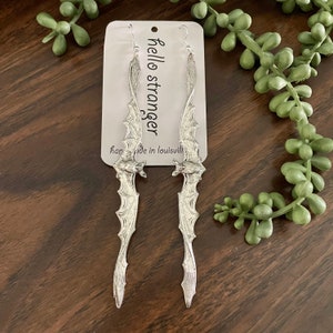 May include: Silver-toned bat wing earrings with a detailed wing design. The earrings hang from silver hooks and are displayed on a white tag with the text "hello stranger" and "handmade in louisville".