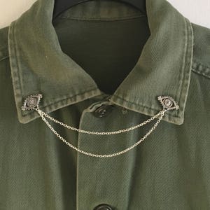 K&ouml;nnte beinhalten: Ein gr&uuml;nes Hemd mit Kragen und zwei silbernen, augenf&ouml;rmigen Anstecknadeln, die durch eine silberne Kette verbunden sind.