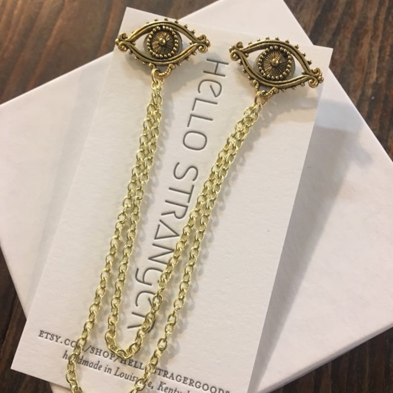 May include: A pair of gold eye charm earrings with a chain drop. The earrings are on a white card with the text "HELLO STRANGE" printed on it.