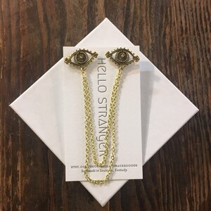 May include: Two gold eye-shaped charms with a chain attached to each. The charms are on a white card with the text "HELLO STRANGER" and "ETSY.COM/SHOP/BELLATRAGERGOODS handmade in Louisville, Kentucky".
