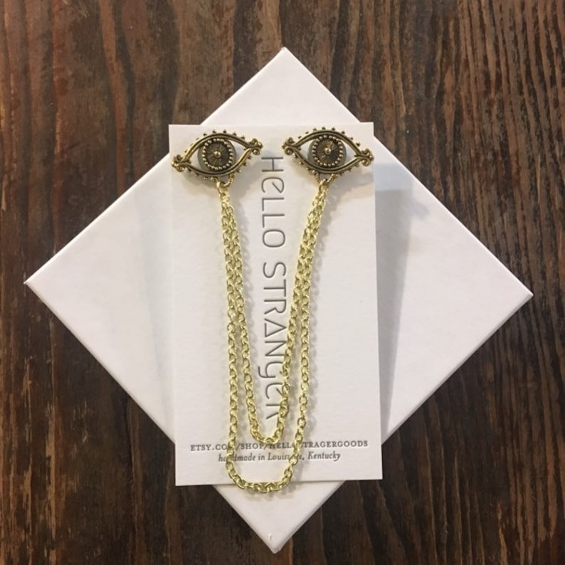 May include: Two gold eye-shaped charms with a chain attached to each. The charms are on a white card with the text "HELLO STRANGER" and "ETSY.COM/SHOP/BELLATRAGERGOODS handmade in Louisville, Kentucky".