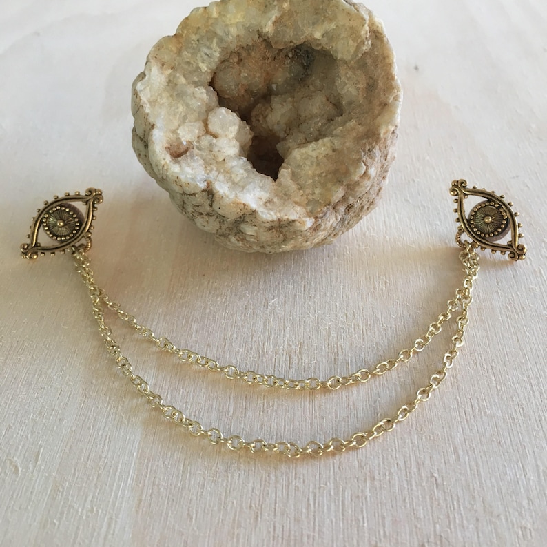 May include: A pair of gold-toned eye charm earrings with a chain connecting them. The earrings are on a white wooden surface.