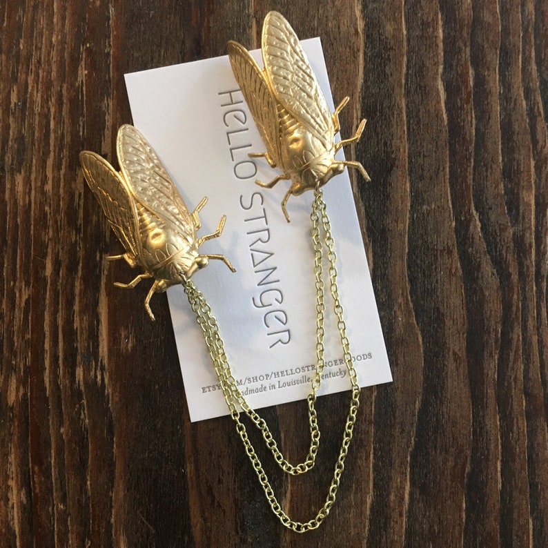 May include: Two gold cicada brooches with chains hanging from them. The brooches are on a white card with the text "HELLO STRANGER" printed on it. The card is on a wooden surface.
