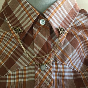 May include: A gold collar pin with a chain and two eye-shaped charms. The pin is attached to a brown and white plaid shirt.