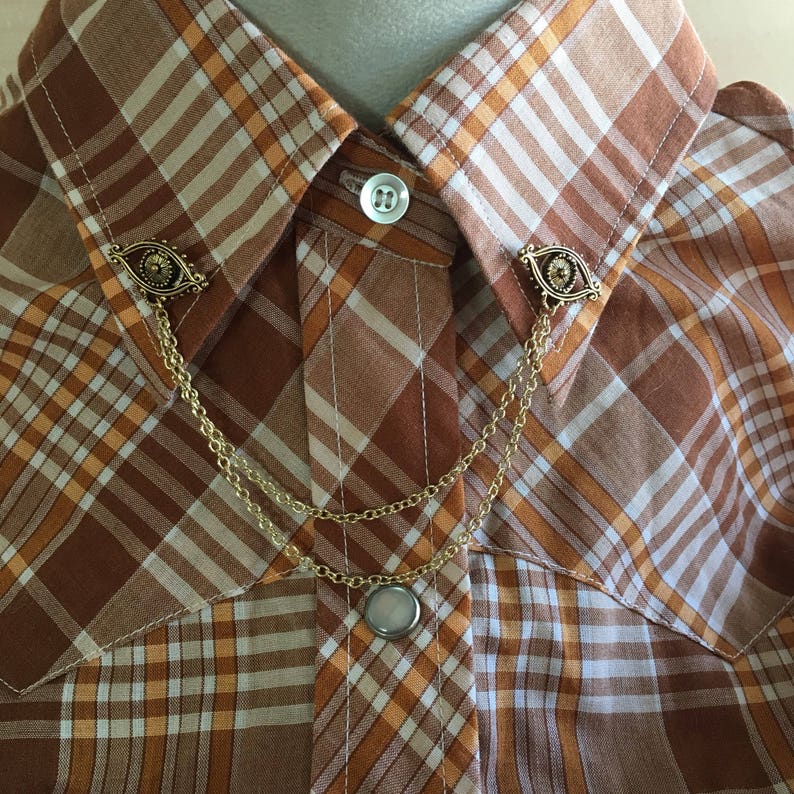May include: A gold collar pin with a chain and two eye-shaped charms. The pin is attached to a brown and white plaid shirt.