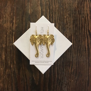 May include: Gold-colored elephant earrings with detailed trunk and ear features. The earrings are displayed on a white card inside a white box, set against a dark wood grain background. The card has the text "HELLO STRANGER" and a website address.