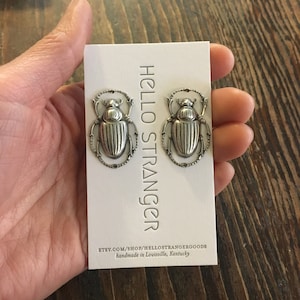 May include: A pair of silver-toned beetle stud earrings on a white card with the text "HELLO STRANGER". The earrings are detailed with textured wings and a decorative border. The card also includes the Etsy shop URL and location.