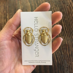 May include: A pair of gold-colored scarab beetle earrings on a white card. The card has the text "HELLO STRANGER" and the Etsy shop URL. The earrings are detailed with intricate designs, and the card is held by a hand.
