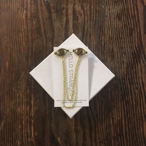 May include: A pair of gold eye charm earrings with a chain hanging from each charm. The earrings are on a white card with the text "HELLO STRANGER" and the website address "etsy.com/shop/hellostrangergoods".