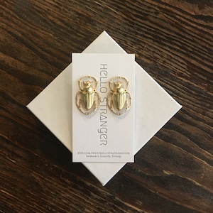 May include: Gold-colored scarab beetle earrings on a white card with the text "HELLO STRANGER". The earrings are displayed on a white box, set against a dark wood grain surface. The earrings are a unique jewelry item.