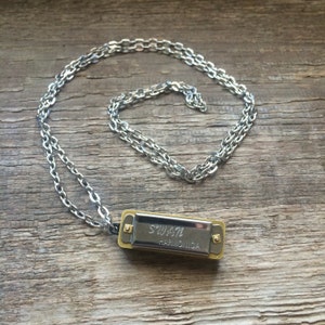 May include: A silver chain necklace with a small harmonica pendant. The harmonica is silver and gold and has the text "SWATI HARMONICA" on it.