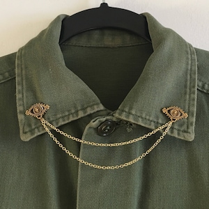May include: A green collared shirt with two gold eye-shaped pins connected by a gold chain.
