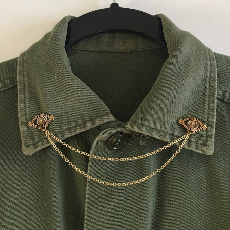 May include: A green collared shirt with two gold eye-shaped pins connected by a gold chain.