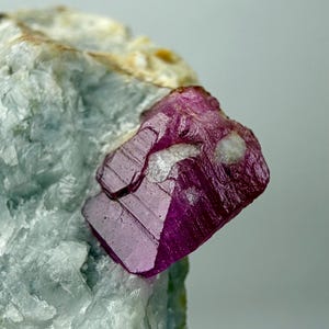 Puede incluir: Un cristal en bruto de color magenta intenso incrustado en una matriz mineral texturizada de color gris pálido y blanco. El cristal presenta facetas angulares afiladas y una superficie translúcida y brillante con inclusiones internas.