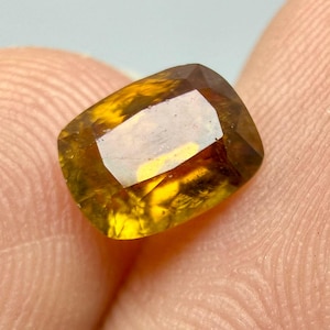 Puede incluir: Una gema facetada, de corte rectangular, con un cálido tono amarillo dorado. La gema tiene una superficie pulida que refleja la luz, mostrando su estructura interna y claridad. Los bordes de la piedra son de un color marrón-anaranjado más oscuro.