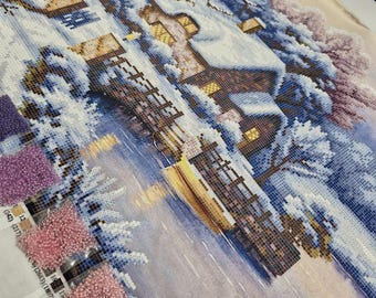 May include: A winter scene with a house, trees, and a river. The image is made up of small, colorful beads. The beads are arranged in a grid pattern to create the image. There are several bags of beads in the lower left corner of the image.