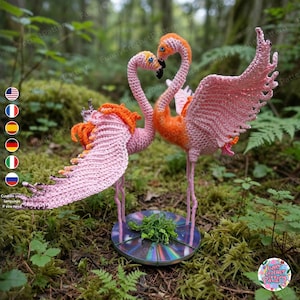 Puede incluir: Dos figuras de flamencos tejidas a ganchillo con cuerpos de hilo rosa y naranja, cuellos largos y alas extendidas. Los flamencos se apoyan sobre una base circular reflectante con un pequeño grupo de material verde en el centro.