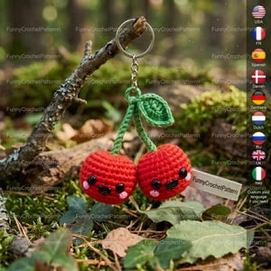 Può includere: Un portachiavi a forma di ciliegie rosse all'uncinetto, con gambo e foglia verdi. Le ciliegie hanno occhi neri e guance rosa. Il portachiavi è appeso a un anello d'argento. Lo sfondo è un ambiente naturale sfocato.