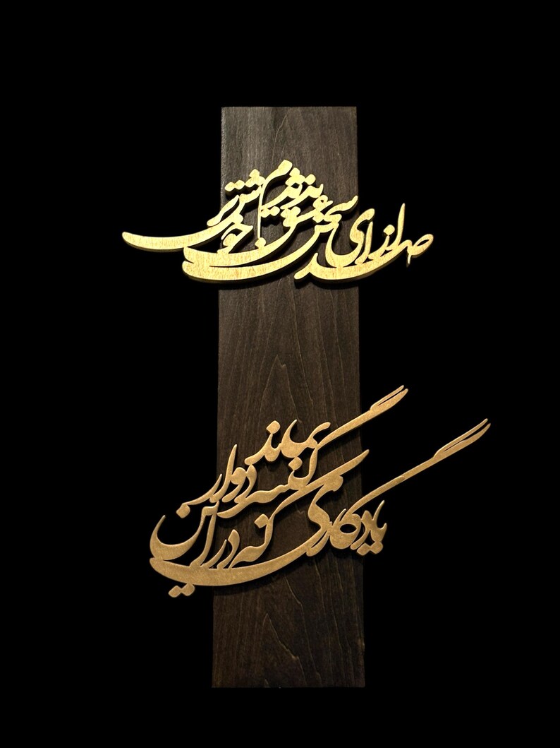 May include: A wooden plaque with two gold-colored, ornate calligraphic designs. The plaque is dark brown, and the background is black. The calligraphy appears to be in a script style, with flowing lines and curves.