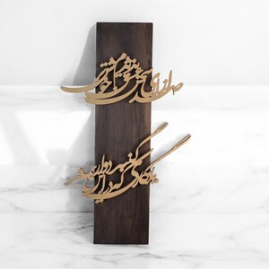 May include: A dark brown wooden wall hanging with gold-colored calligraphy. The calligraphy is in two sections, one above the other. The background is a white marble surface.