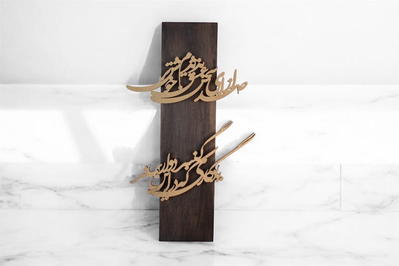 May include: A dark brown wooden wall hanging with gold-colored calligraphy. The calligraphy is in two sections, one above the other. The background is a white marble surface.