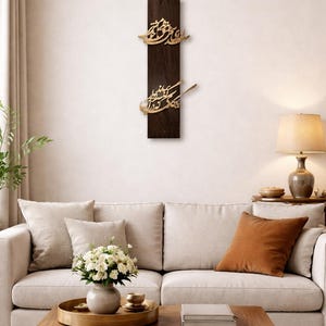 May include: A dark brown wooden wall art piece with gold-colored Arabic calligraphy. The artwork is rectangular and mounted on a white wall above a sofa. The room is decorated with neutral tones and a wooden coffee table.