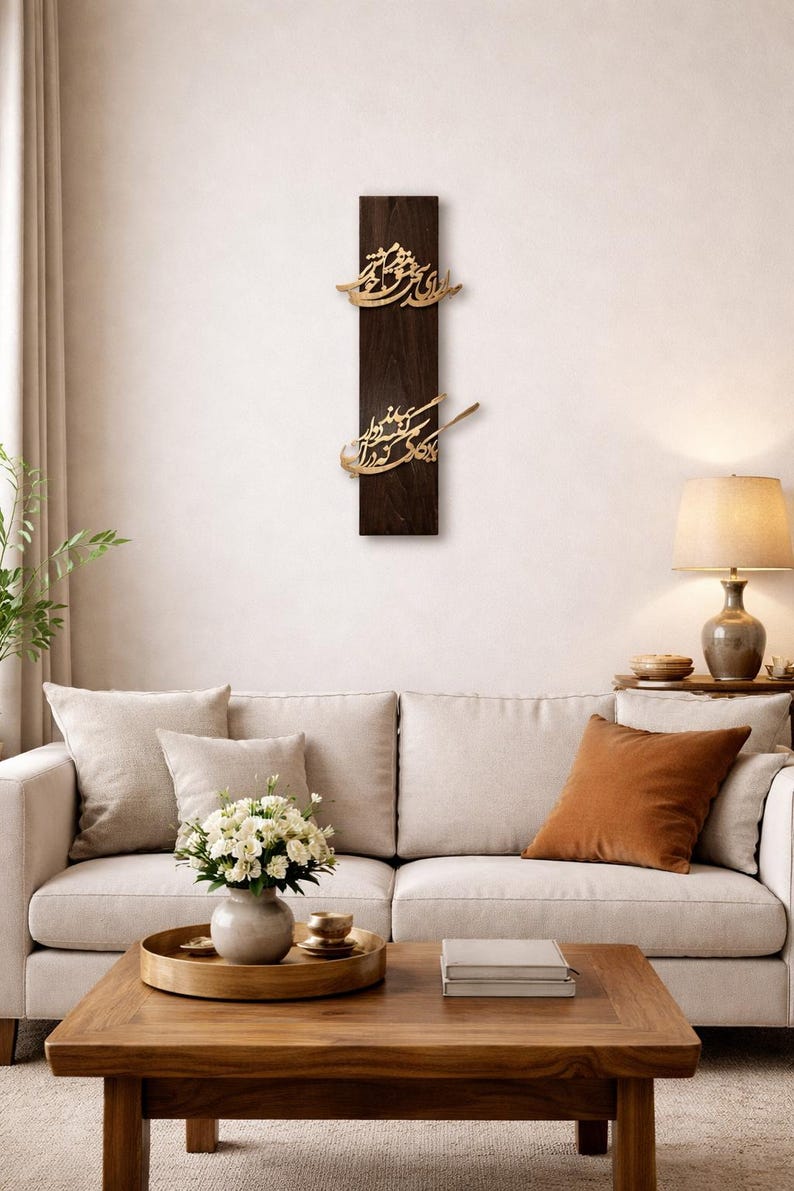 May include: A dark brown wooden wall art piece with gold-colored Arabic calligraphy. The artwork is rectangular and mounted on a white wall above a sofa. The room is decorated with neutral tones and a wooden coffee table.