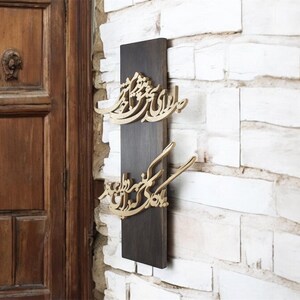 May include: A dark brown wooden wall hanging with gold-colored Arabic calligraphy. The calligraphy is mounted on a rectangular dark brown wooden board, against a white brick wall. The door in the background is made of wood.