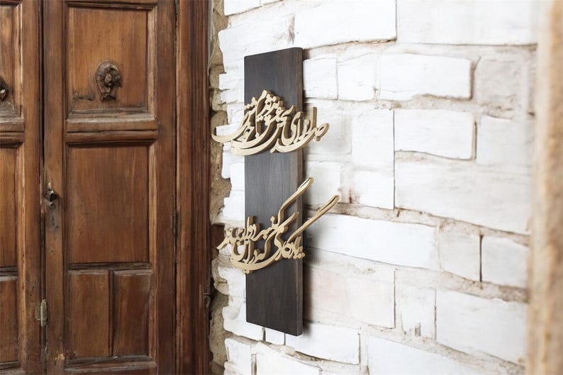 May include: A dark brown wooden wall hanging with gold-colored Arabic calligraphy. The calligraphy is mounted on a rectangular dark brown wooden board, against a white brick wall. The door in the background is made of wood.