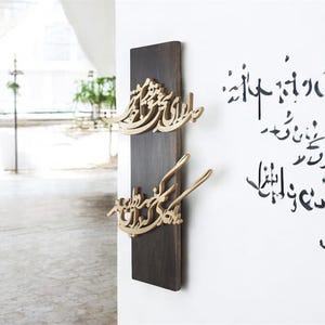 May include: A dark brown wooden wall hanging with gold-colored Arabic calligraphy. The calligraphy is in a flowing script and is mounted on a rectangular wooden plaque. Additional calligraphy is written on the white wall.