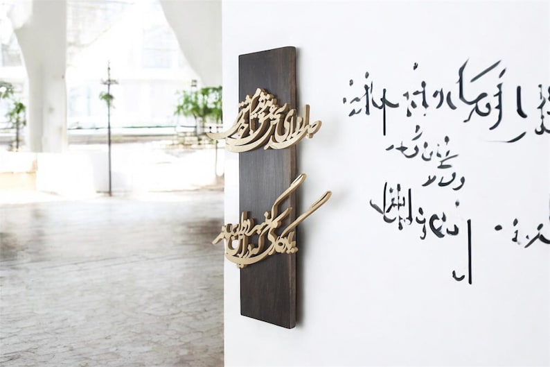 May include: A dark brown wooden wall hanging with gold-colored Arabic calligraphy. The calligraphy is in a flowing script and is mounted on a rectangular wooden plaque. Additional calligraphy is written on the white wall.
