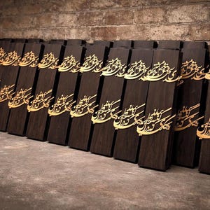 May include: A collection of dark brown wooden plaques with gold-colored calligraphy. The plaques are rectangular and arranged in a row, leaning against a brick wall. The calligraphy appears to be an ornate script.