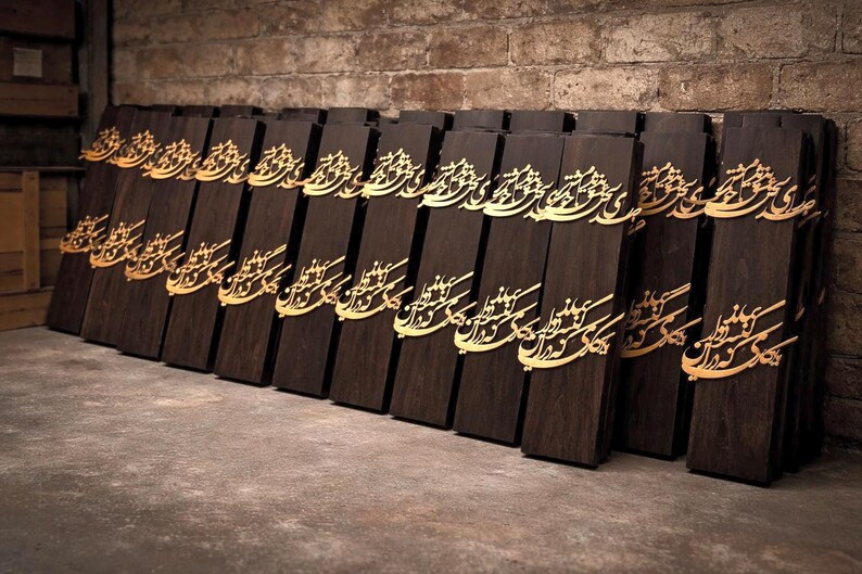 May include: A collection of dark brown wooden plaques with gold-colored calligraphy. The plaques are rectangular and arranged in a row, leaning against a brick wall. The calligraphy appears to be an ornate script.