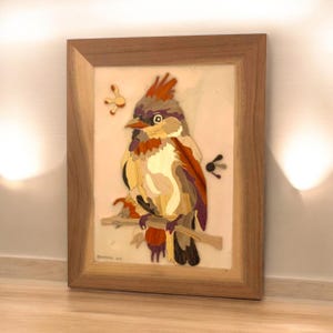 May include: A framed wood marquetry artwork depicting a colorful bird perched on a branch. The bird is made of various wood pieces, creating a mosaic effect. The background is a light pink.