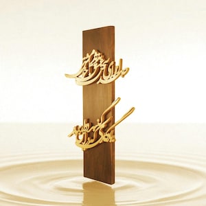 May include: A vertical wooden plaque with gold-colored Arabic calligraphy. The plaque is a dark brown color and stands upright against a light background. The calligraphy is ornate and detailed.
